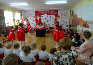 Na tle dekoracji z mapą Polski i dziećmi z chorągiewkami przedszkolaki wykonują układ taneczny do utworu "Prząśniczki".